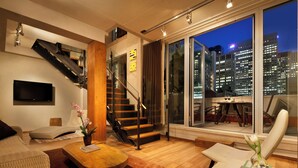 Una sala de estar moderna con una escalera, un televisor de pantalla plana, una mesa de centro y vistas a un paisaje urbano nocturno.