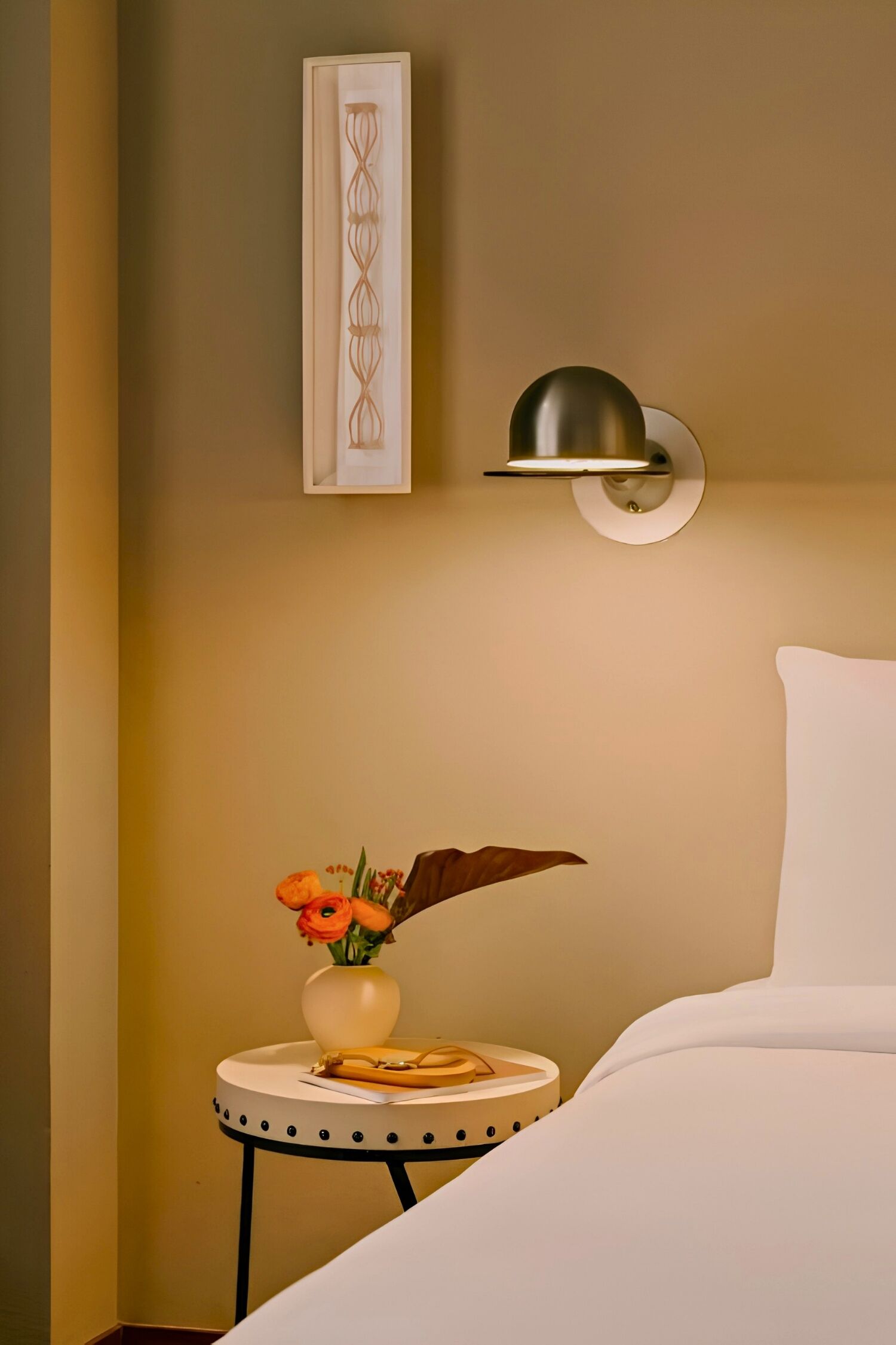 A neatly arranged hotel room with a bed, a bedside table with a vase of flowers, a wall-mounted lamp, and a framed artwork.