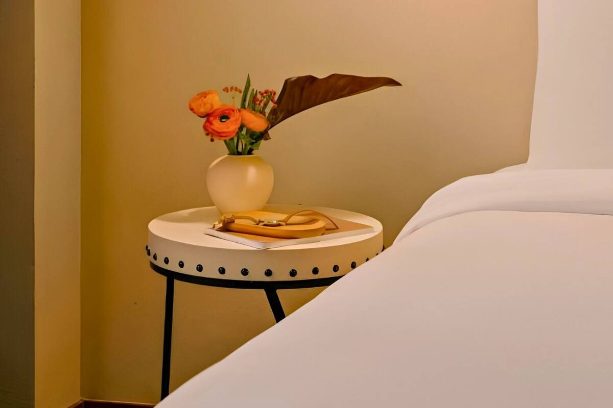Habitación de hotel ordenada con cama, mesita de noche con un florero y una lámpara de pared, además de una obra de arte enmarcada.