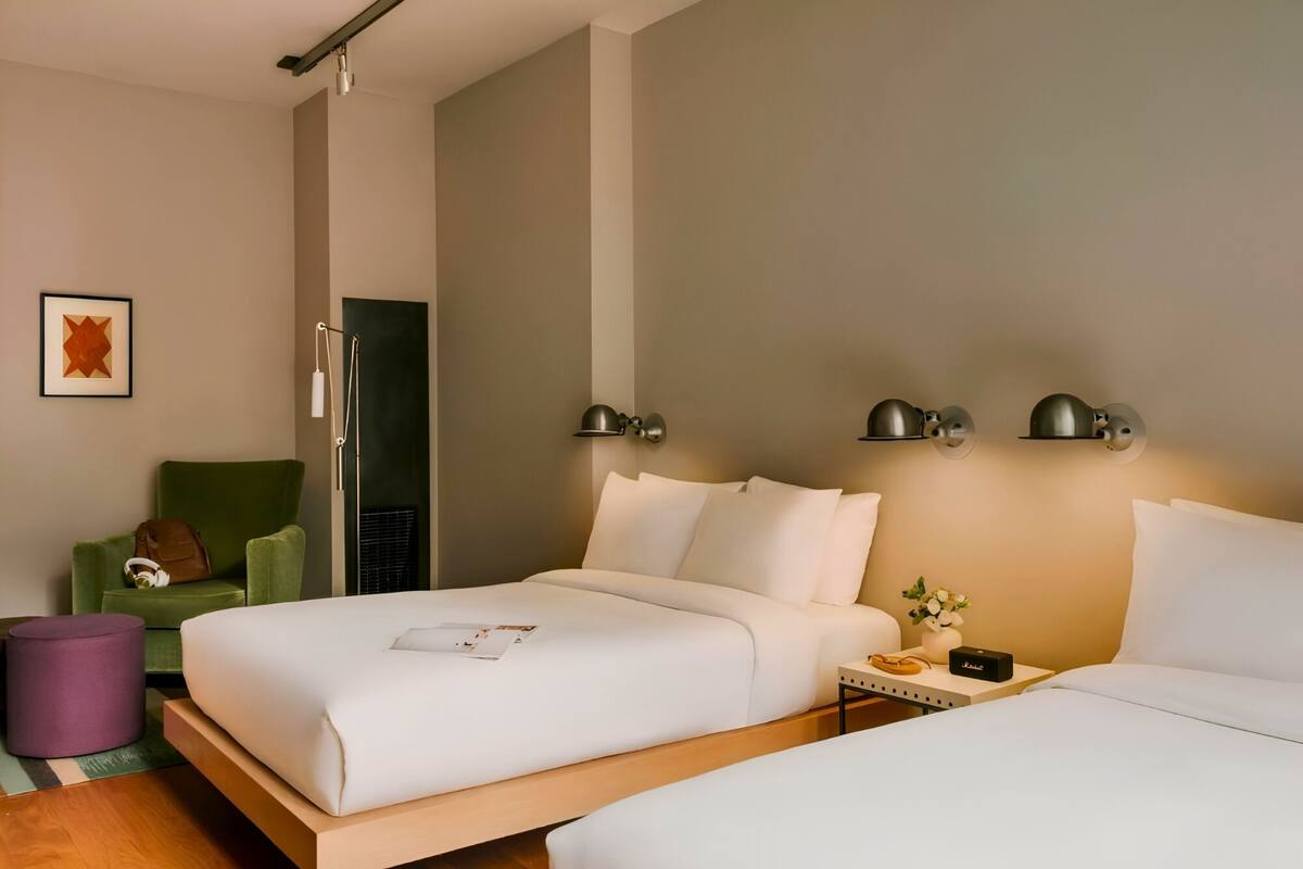 Habitación de hotel con dos camas, un sillón verde, una mesita con un jarrón y un cuadro en la pared.