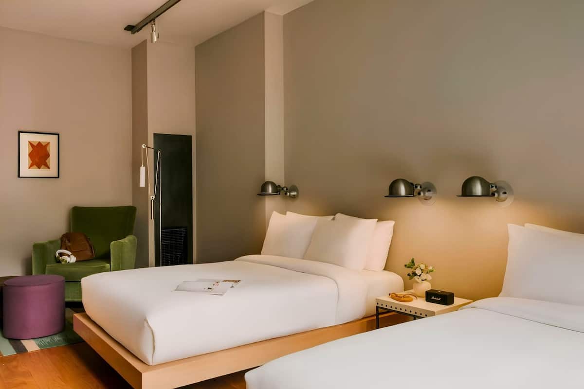 A hotel room with two beds, a green armchair, a small table with a vase, and a framed picture on the wall.