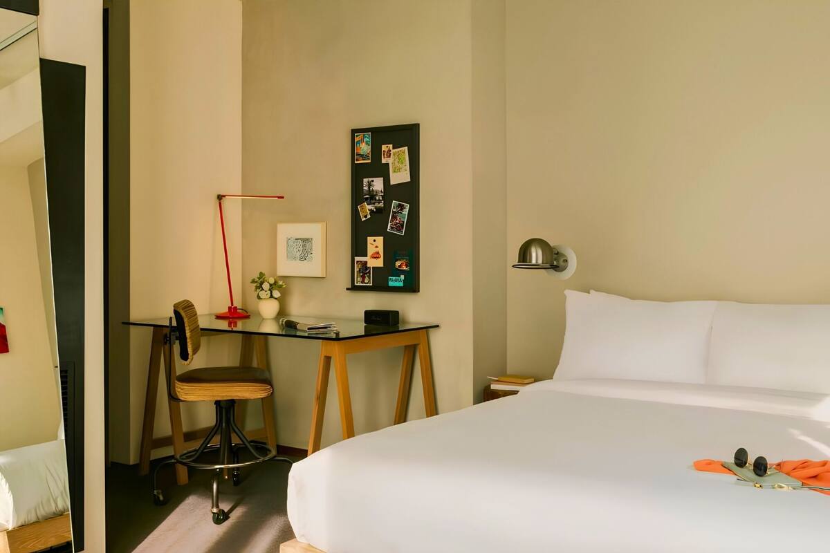 Habitación de hotel con cama, escritorio y silla, lámpara, y pared decorada con fotos y notas.