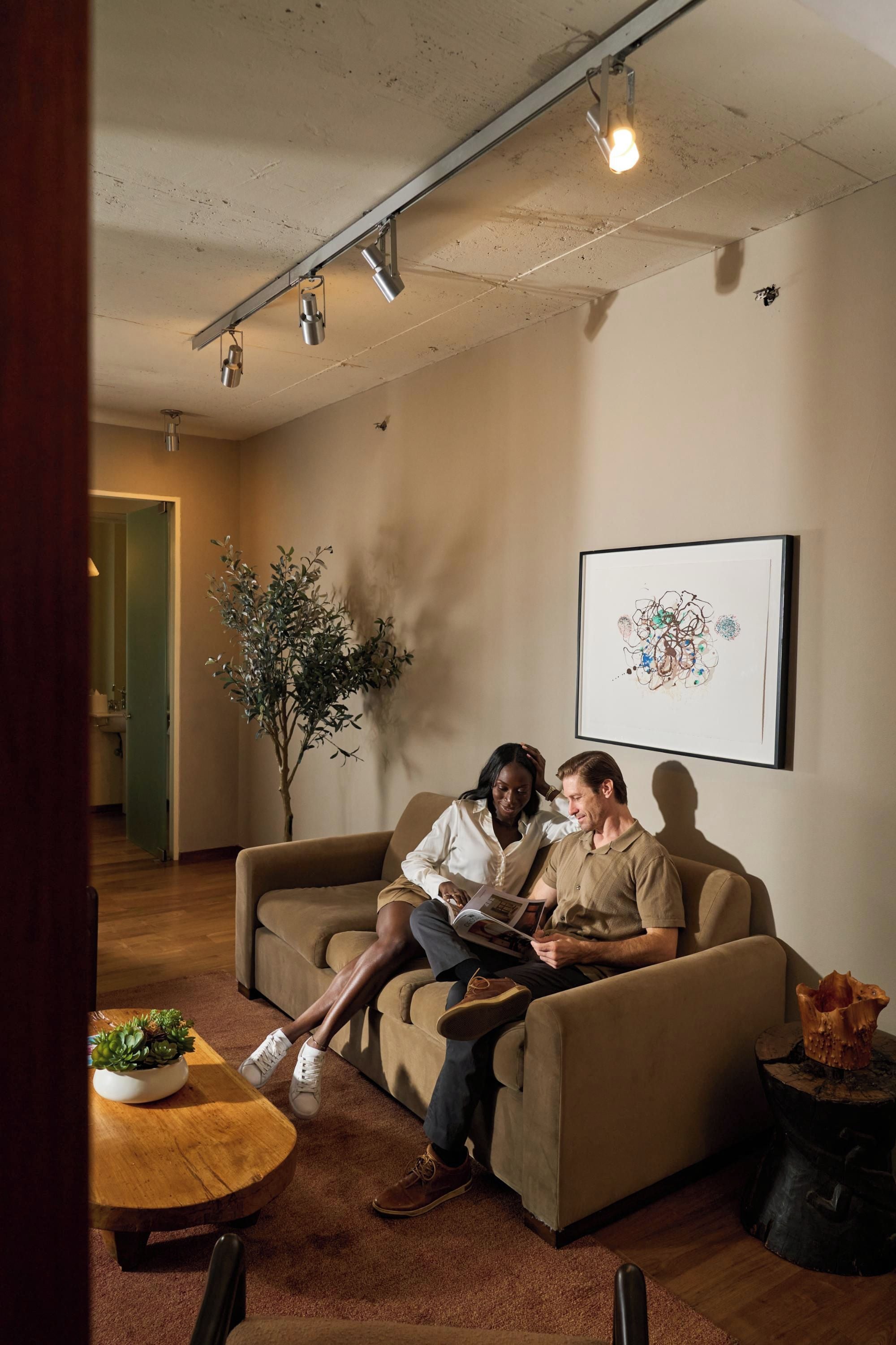 A room with a beige sofa, a coffee table with a plant, a framed abstract painting, and a wooden side table with a decorative item.