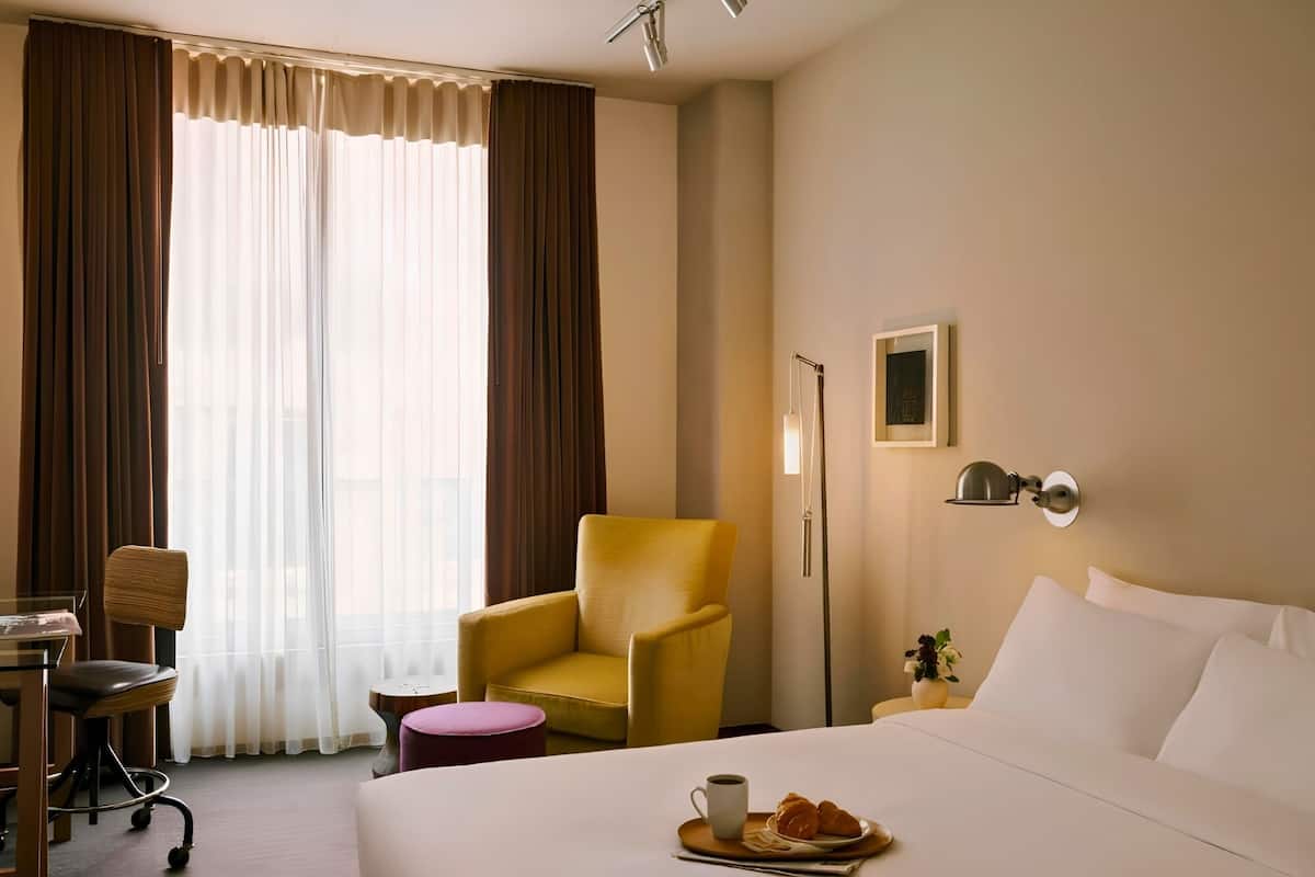 Una habitación de hotel con una cama, un sillón amarillo, un escritorio con una silla, una mesita redonda pequeña y una mesita de noche con una lámpara.