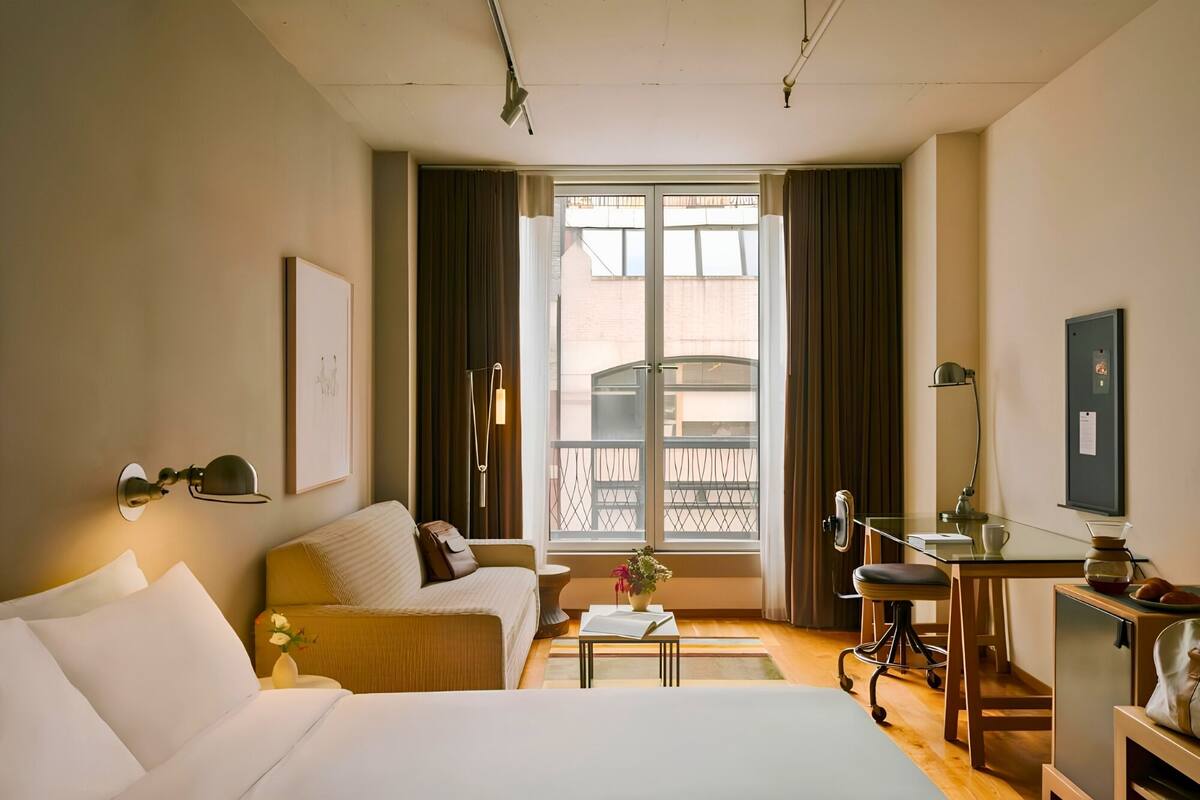 Habitación de hotel con cama, sofá, escritorio, silla, una mesita, una planta en maceta y una ventana con cortinas.