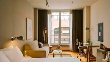 A hotel room with a bed, a sofa, a desk, a chair, a small table, a potted plant, and a window with curtains.