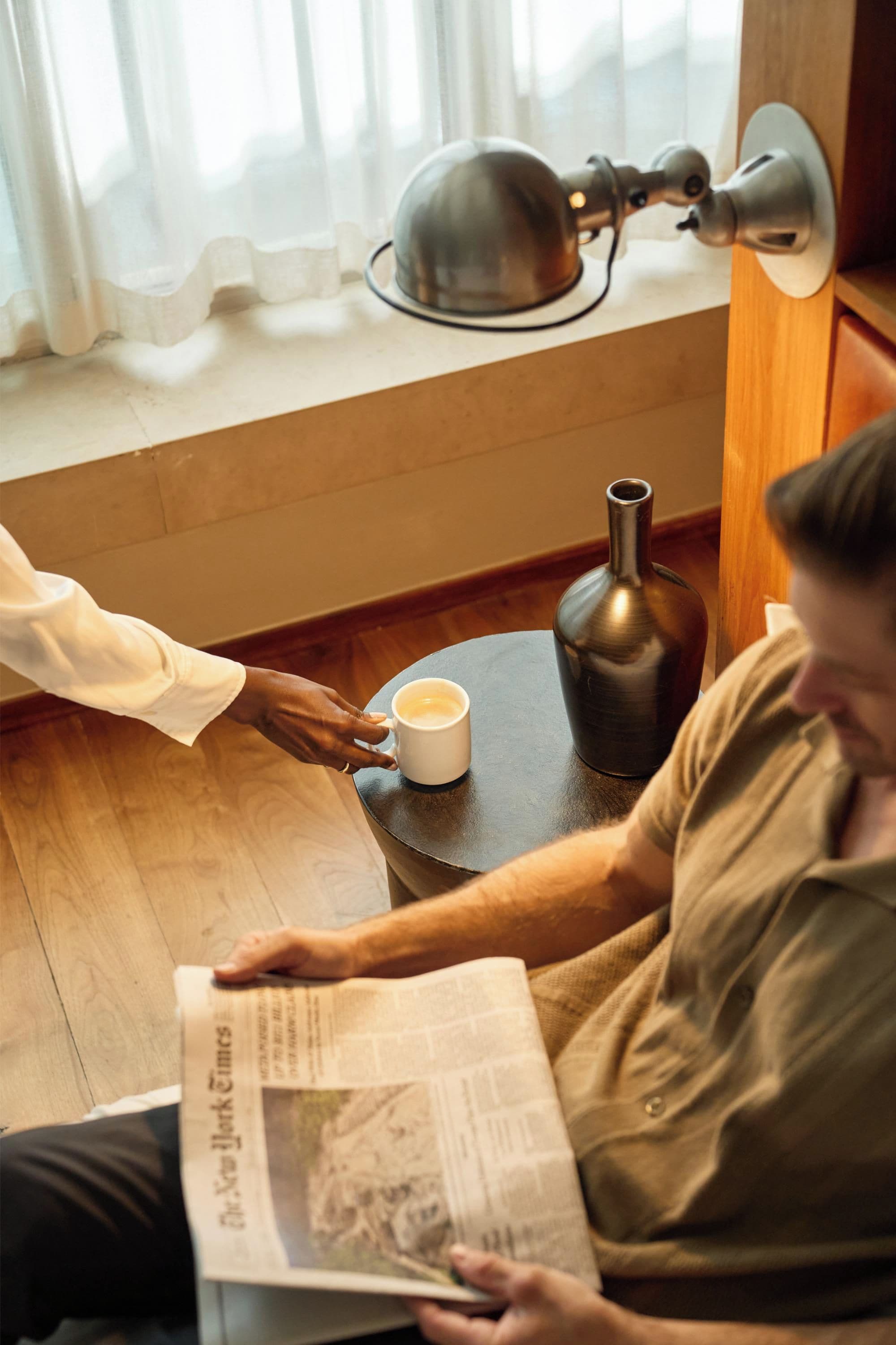 A person sitting on a wooden floor reading a newspaper, with a coffee cup and a lamp on a small table beside them.