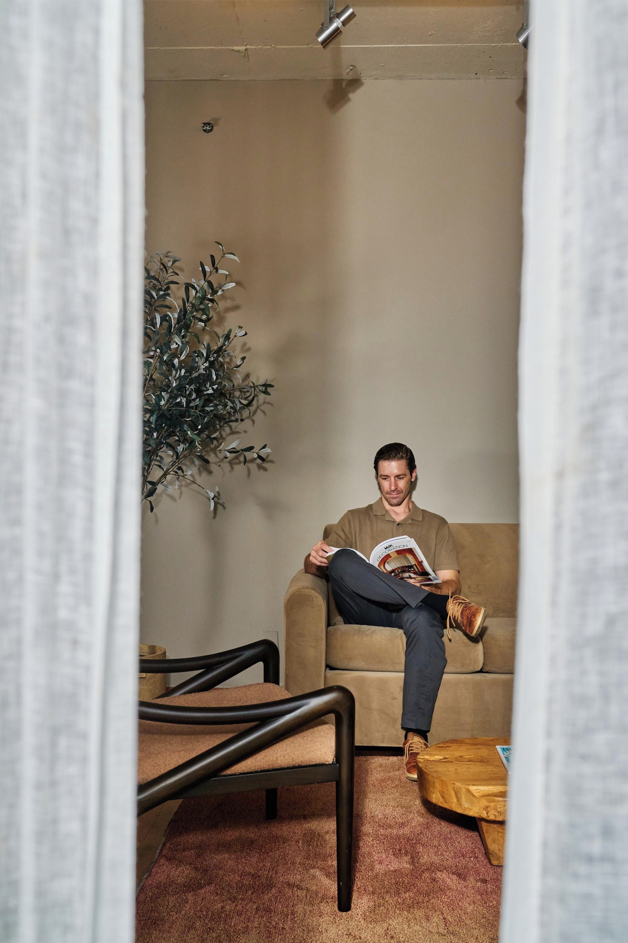 A person sitting on a sofa reading a magazine in a room with a plant, a wooden coffee table, and a chair.