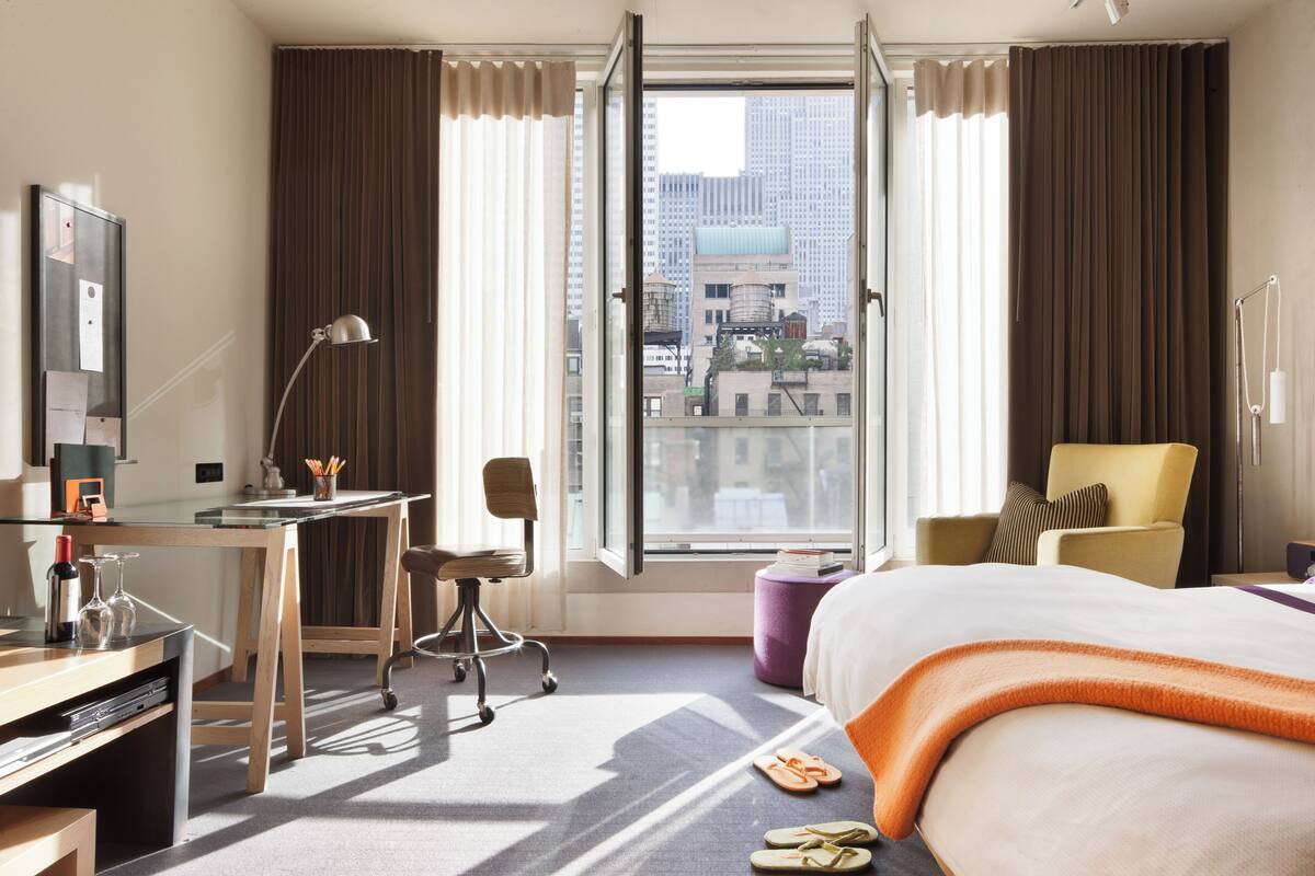 Habitación de hotel con cama, escritorio, silla y una ventana grande con cortinas.