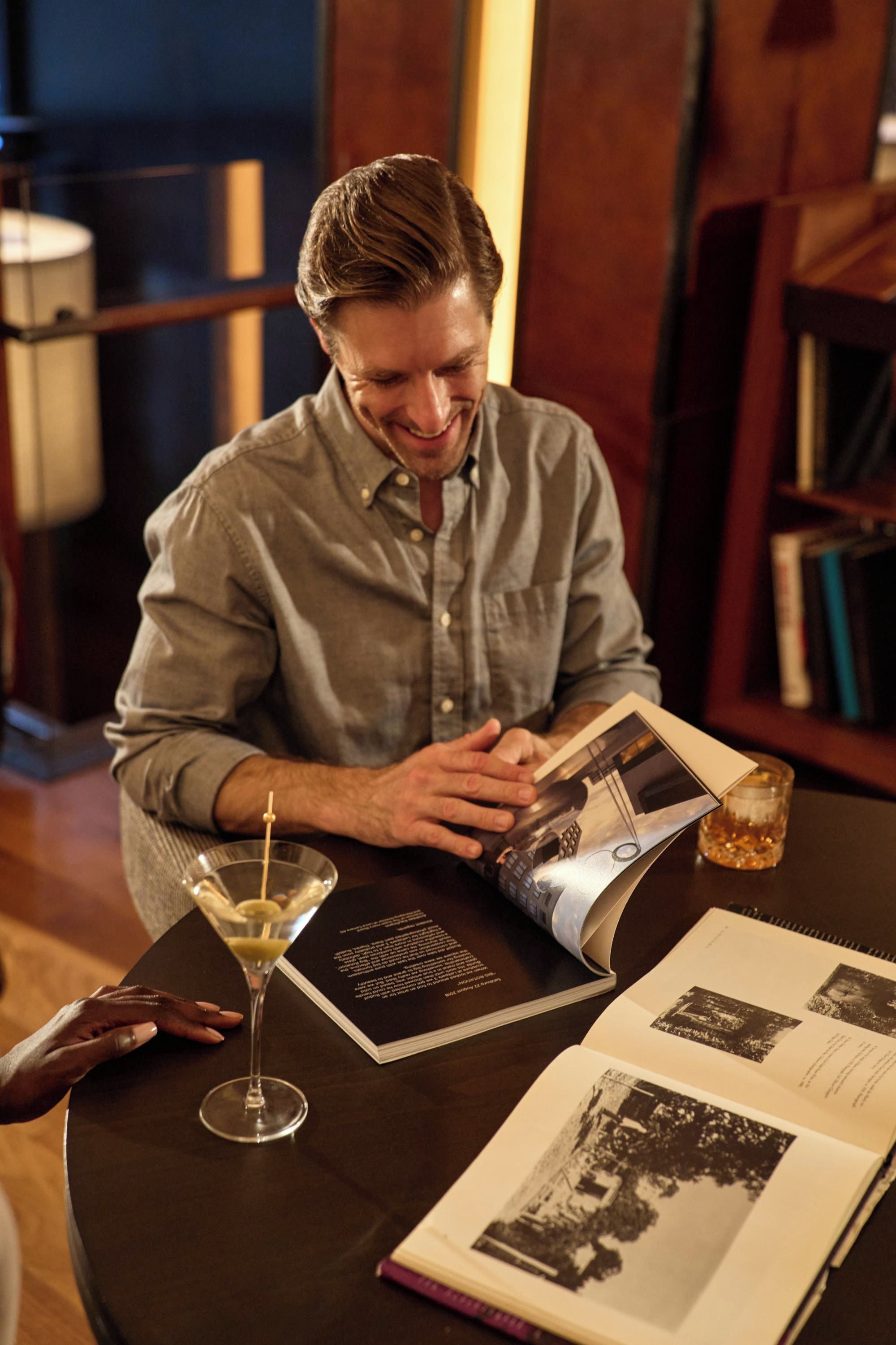 Eine Person liest ein Buch an einem Tisch, auf dem ein Martini und eine Speisekarte stehen.