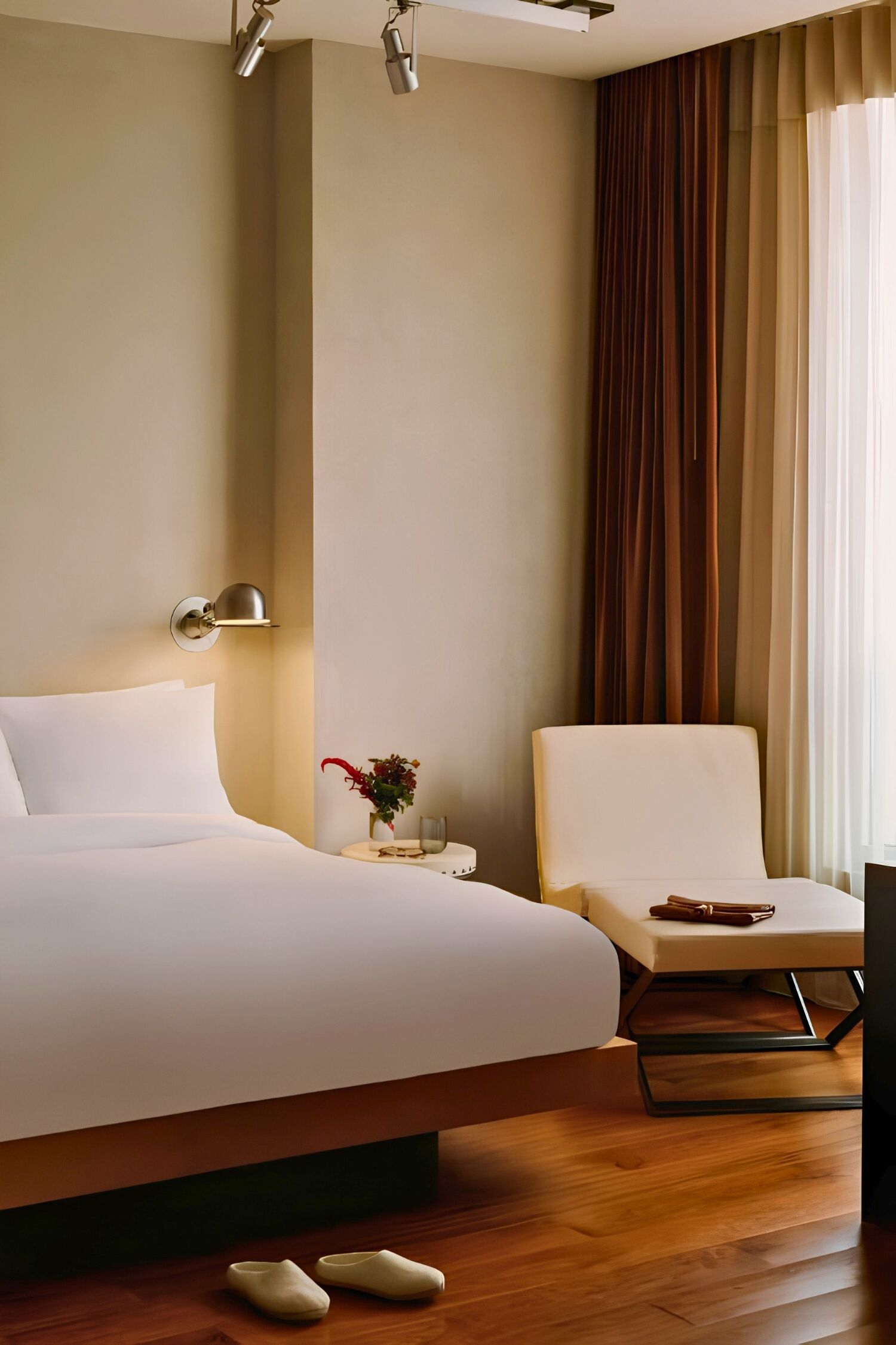 Quarto de hotel com cama, poltrona, mesa de cabeceira com abajur e janela com cortinas.