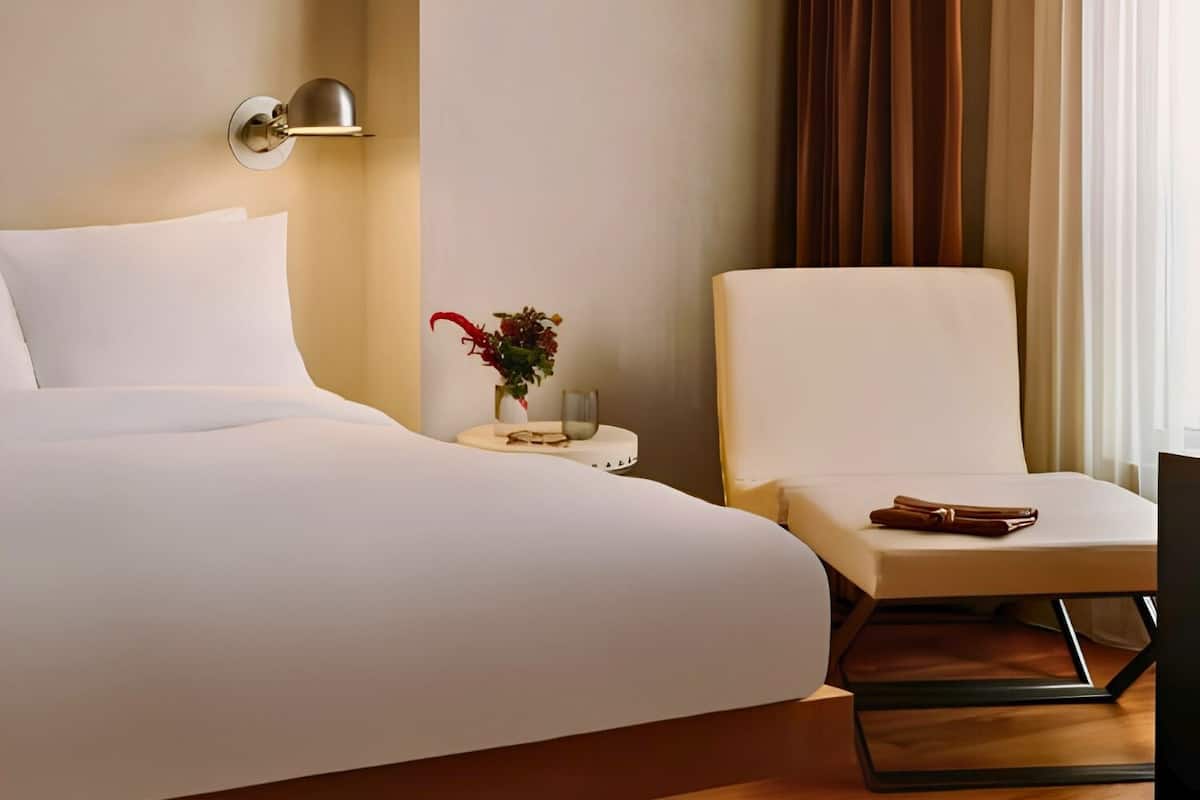 Habitación de hotel con cama, una silla, mesita de noche con lámpara y ventana con cortinas.