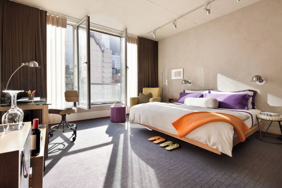 Una habitación de hotel moderna con una cama grande, un escritorio con lámpara, una silla, una mesita y vistas a la ciudad.