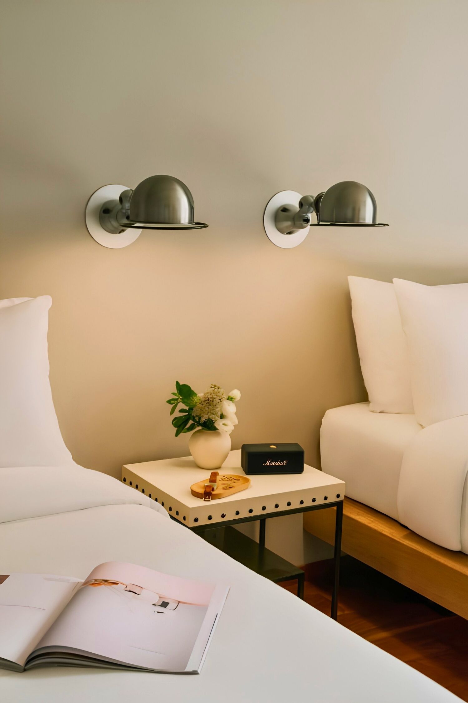 Una habitación de hotel moderna con una cama, una mesita de noche con una lámpara, una pequeña mesa con un jarrón de flores y una revista.