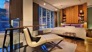 Una habitación de hotel moderna con una cama grande, zona de estar, estantería y vistas a la ciudad.