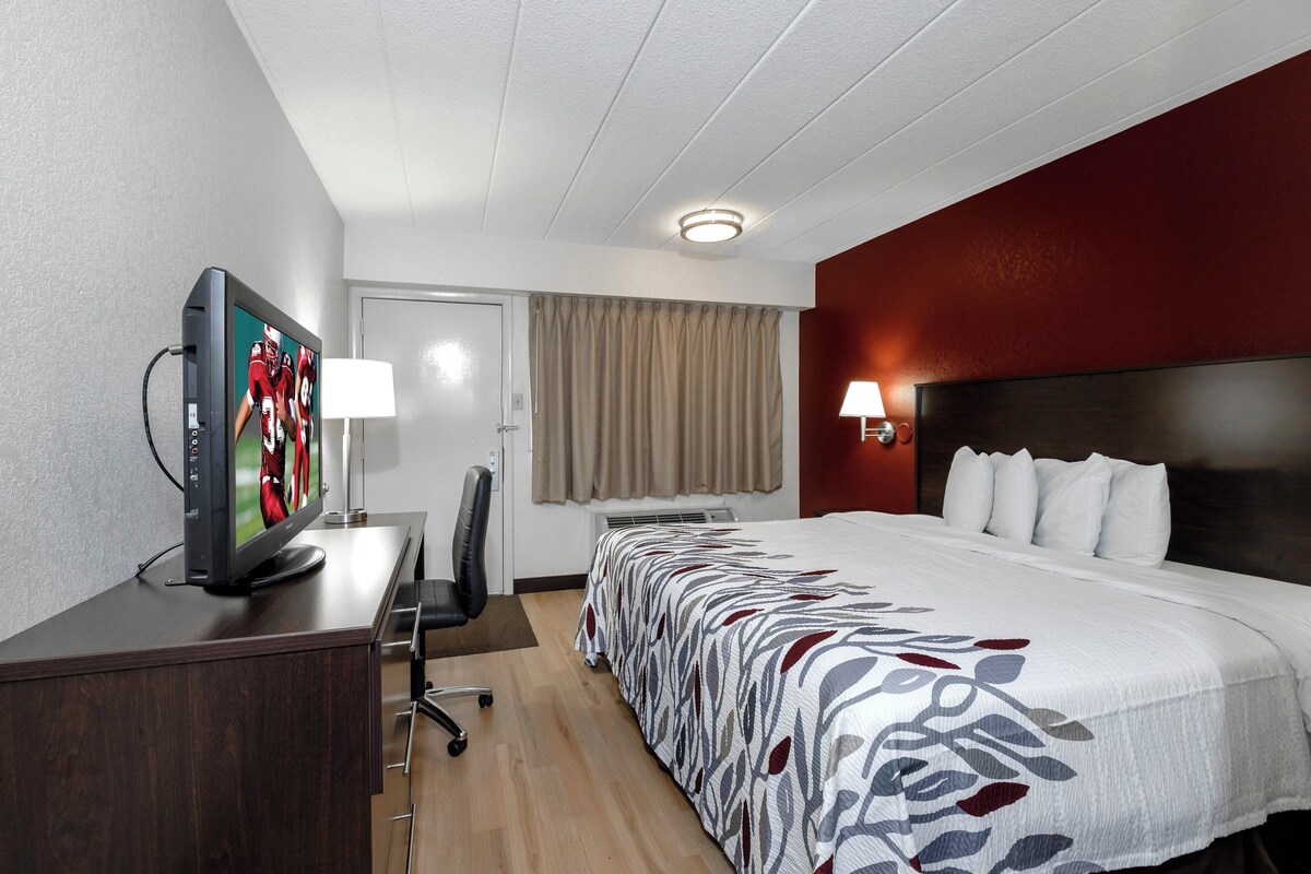 A hotel room with a bed, a desk with a TV, a chair, a lamp, and a window with curtains.