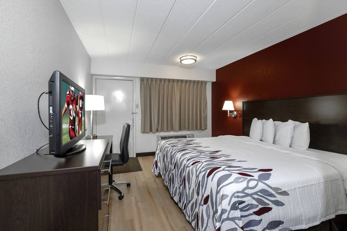 A hotel room with a bed, a desk with a television, a chair, a lamp, and a window with curtains.