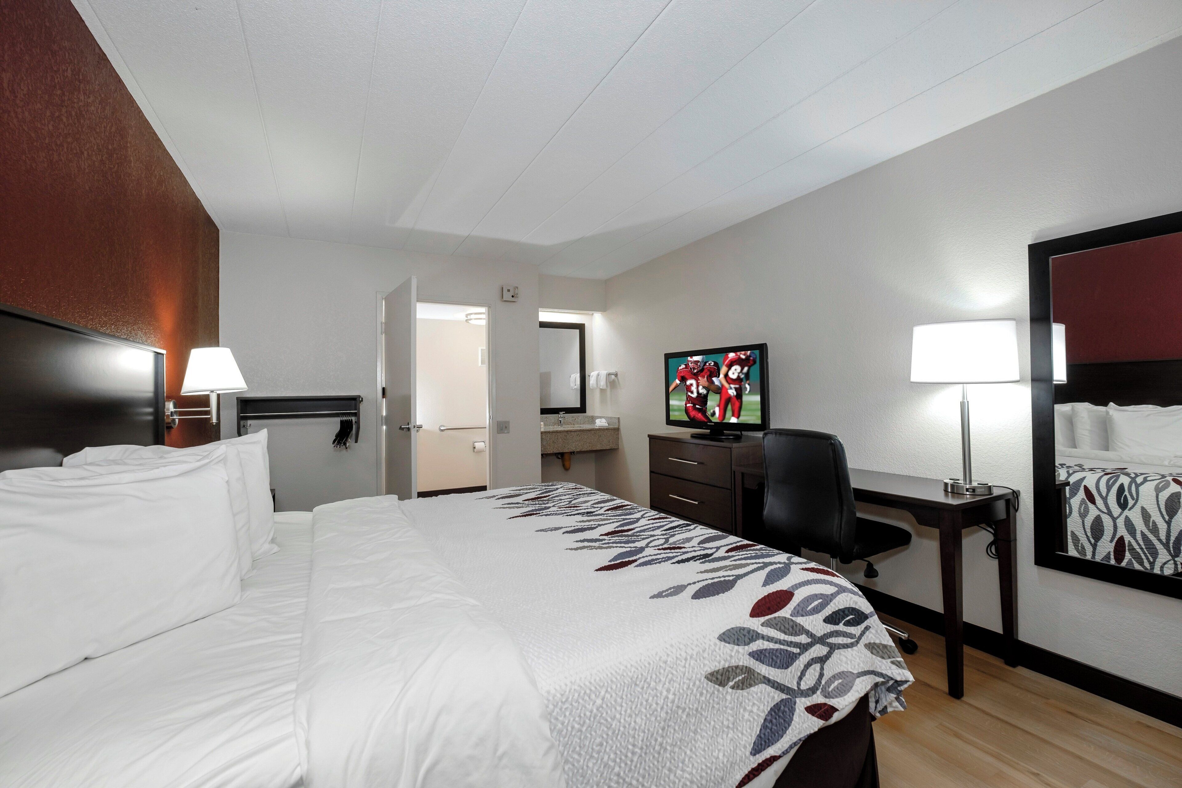 Habitación de hotel con cama, escritorio, silla, televisión y puerta de baño.