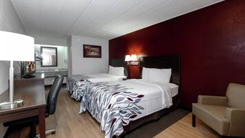 A hotel room with two beds, a desk with a lamp, a chair, and a red accent wall.