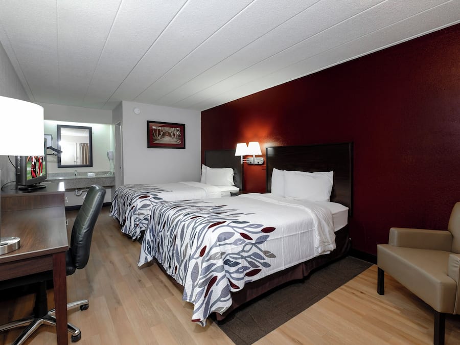 Habitación de hotel con dos camas, un escritorio con lámpara, una silla y una pared de acento roja.