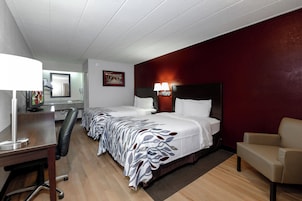 Habitación de hotel con dos camas, un escritorio con lámpara, una silla y una pared de acento roja.