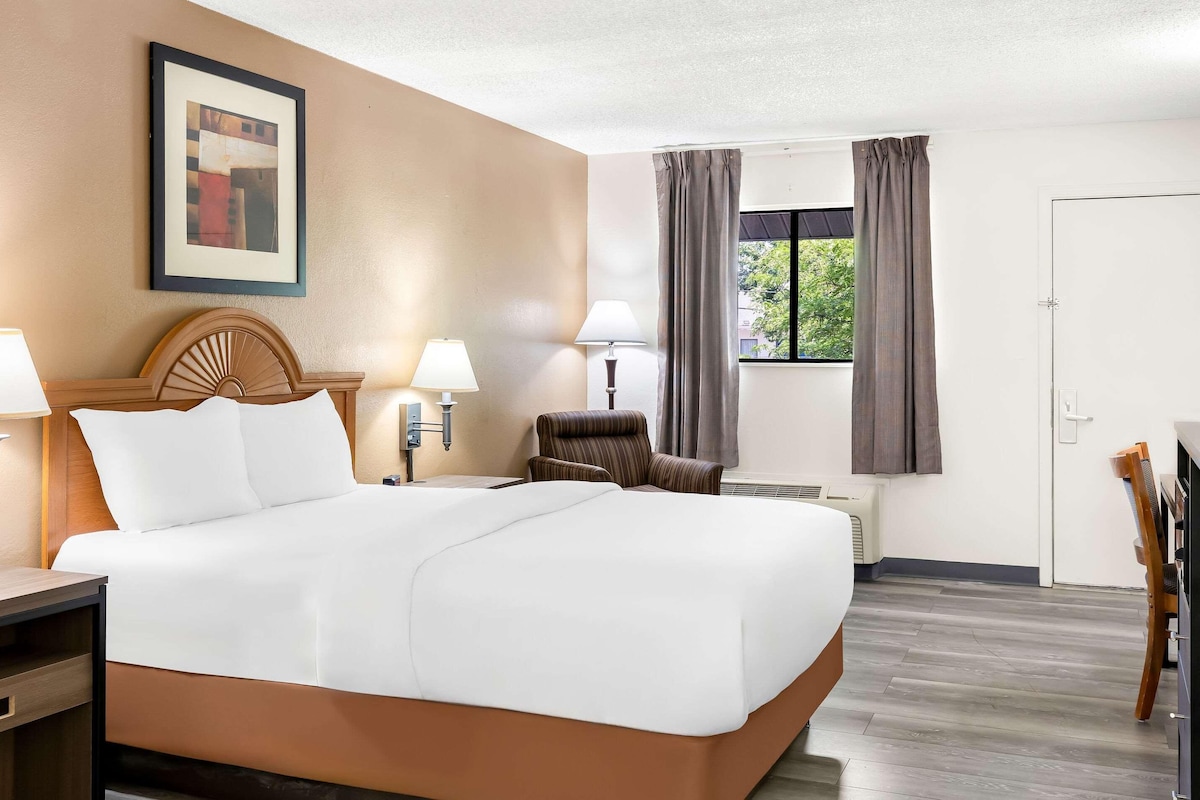 A hotel room with a large bed, two bedside lamps, a chair, a window with curtains, and a framed picture on the wall.