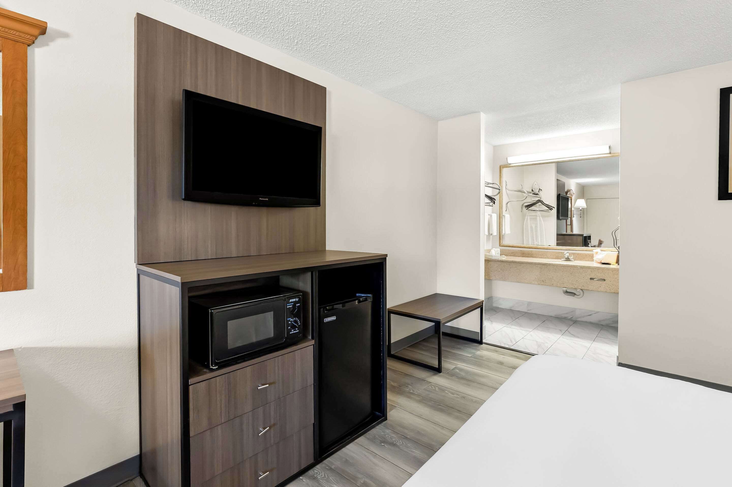 A modern hotel room with a built-in microwave, a small refrigerator, a flat-screen TV mounted on a wooden panel, a minimalist bathroom with a mirror, and a white sink.