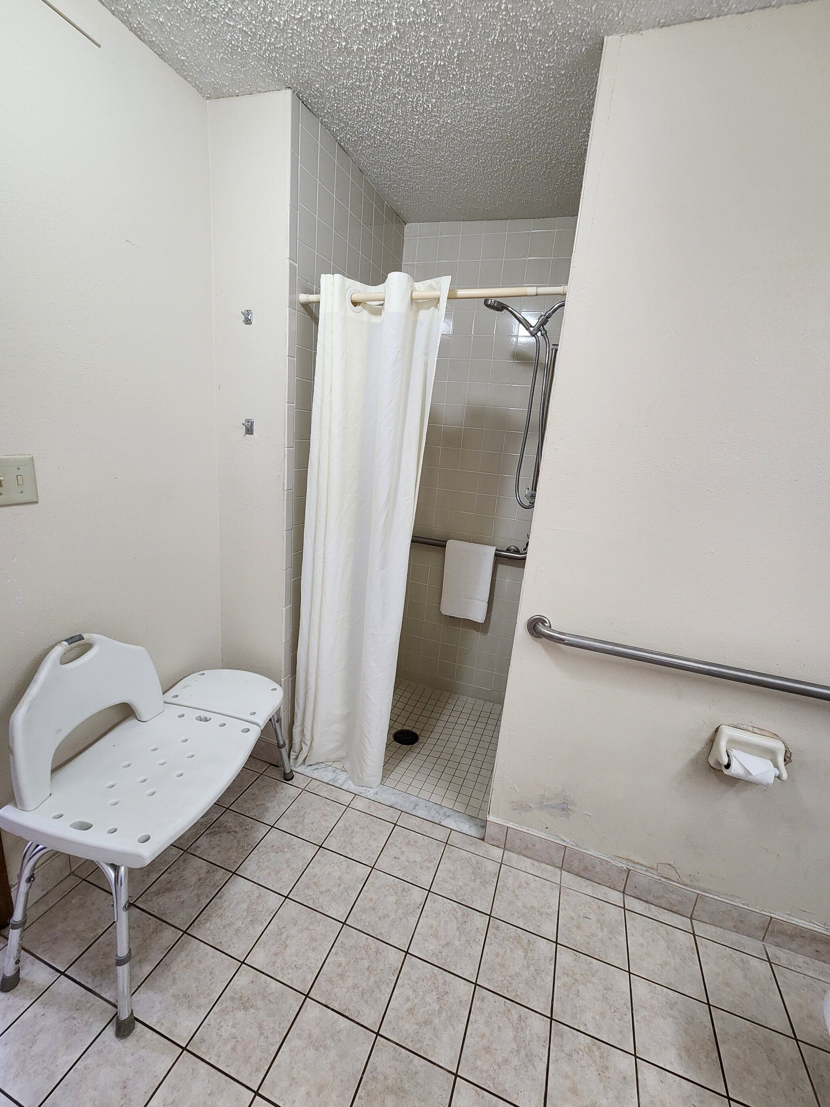 A bathroom with a white shower, a white toilet, and a white bench with armrests.