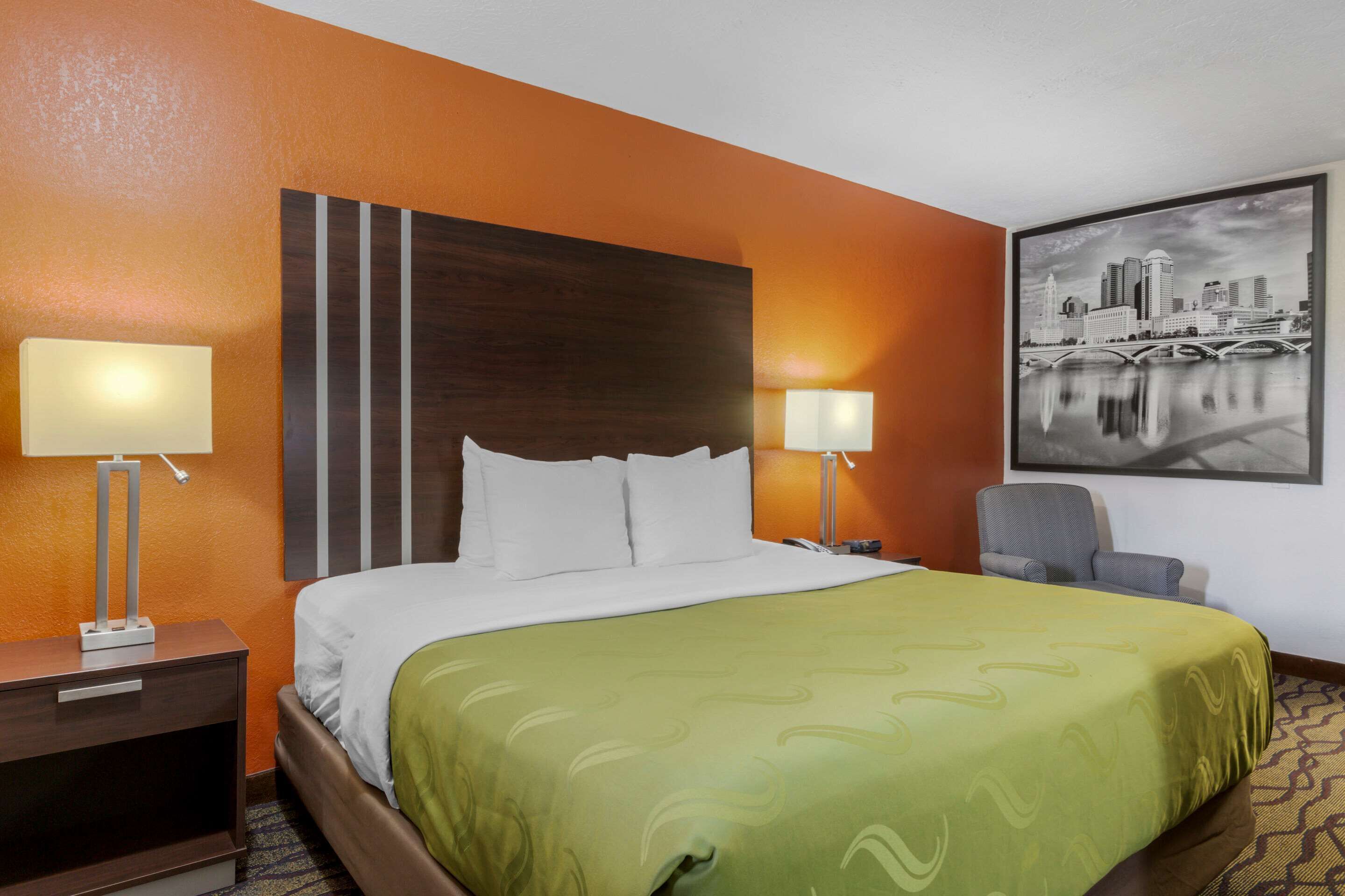 A hotel room with a large bed, two bedside lamps, a chair, and a cityscape painting on the wall.