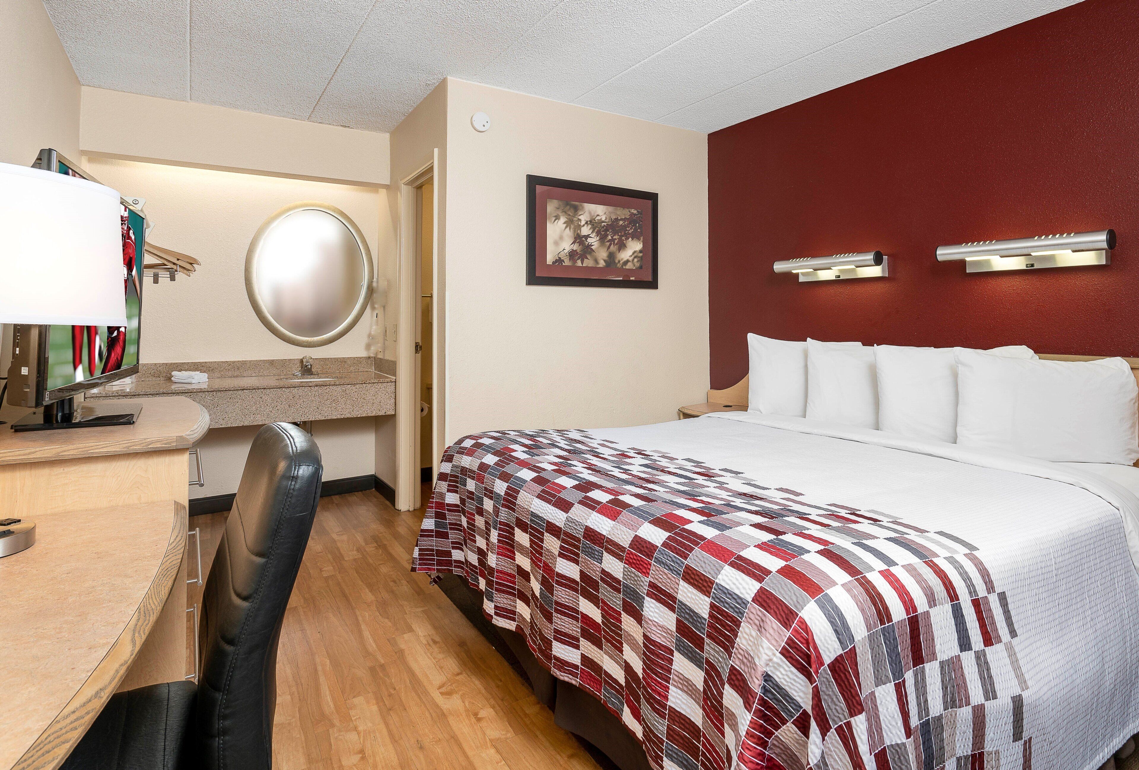 A hotel room with a bed, a desk with a computer, a chair, a mirror, and a wall-mounted light fixture.
