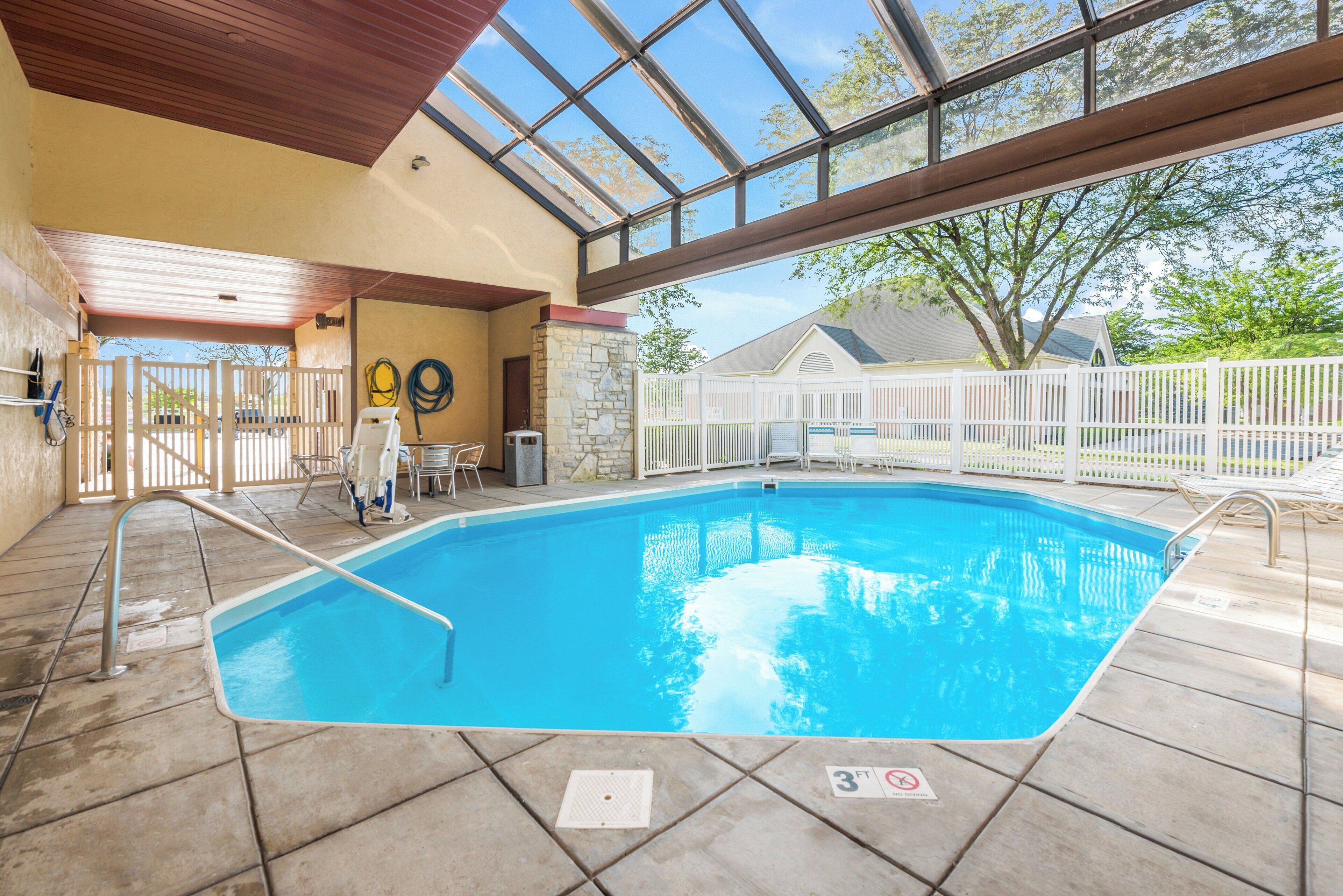 An indoor swimming pool with a clear view of the sky through large windows, surrounded by a white fence and a stone wall.
