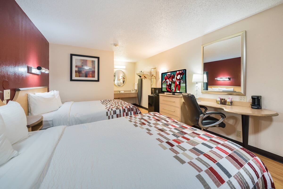 A hotel room with two beds, a desk with a computer, a chair, a mirror, and a TV.