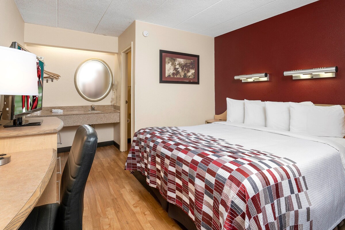 A hotel room with a bed, a desk with a lamp, a chair, a round mirror, and a framed picture on the wall.