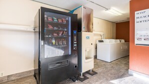Máquina automática com opções de lanches e bebidas, um dispensador de gelo com moedas, e um aviso indicando amaciante de roupa e alvejante.