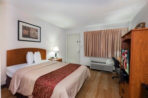 A hotel room with a bed, a desk with a computer, an air conditioner, a lamp, and a framed picture on the wall.