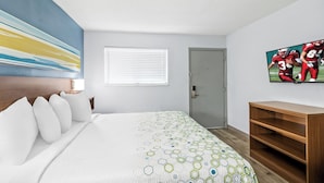 Habitación de hotel con una cama grande, una mesita de noche de madera, un televisor montado en la pared, una puerta y una ventana con persianas.