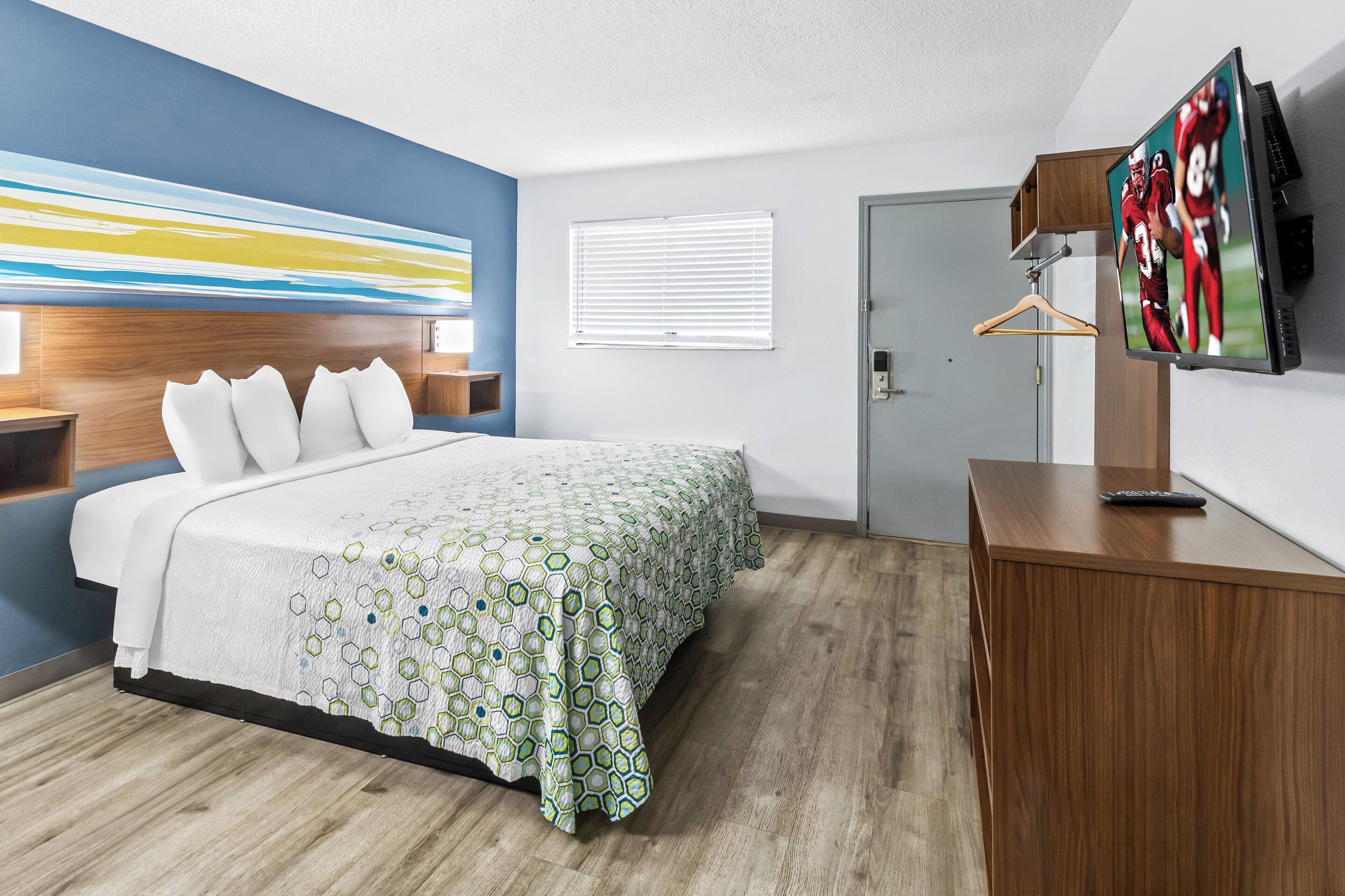 Habitación de hotel con cama, televisor montado en la pared, cómoda de madera y un cuadro colgado.