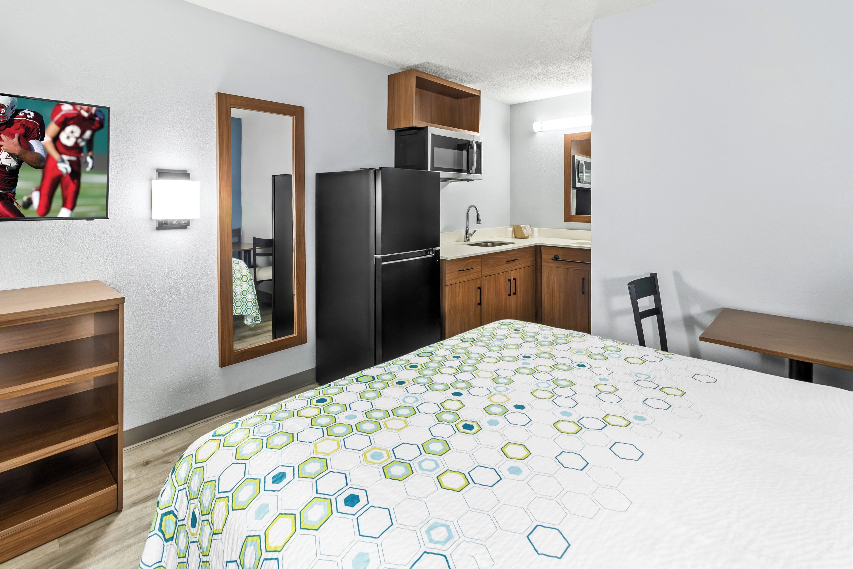 A hotel room with a bed, a desk, a chair, a refrigerator, a microwave, and a sink.