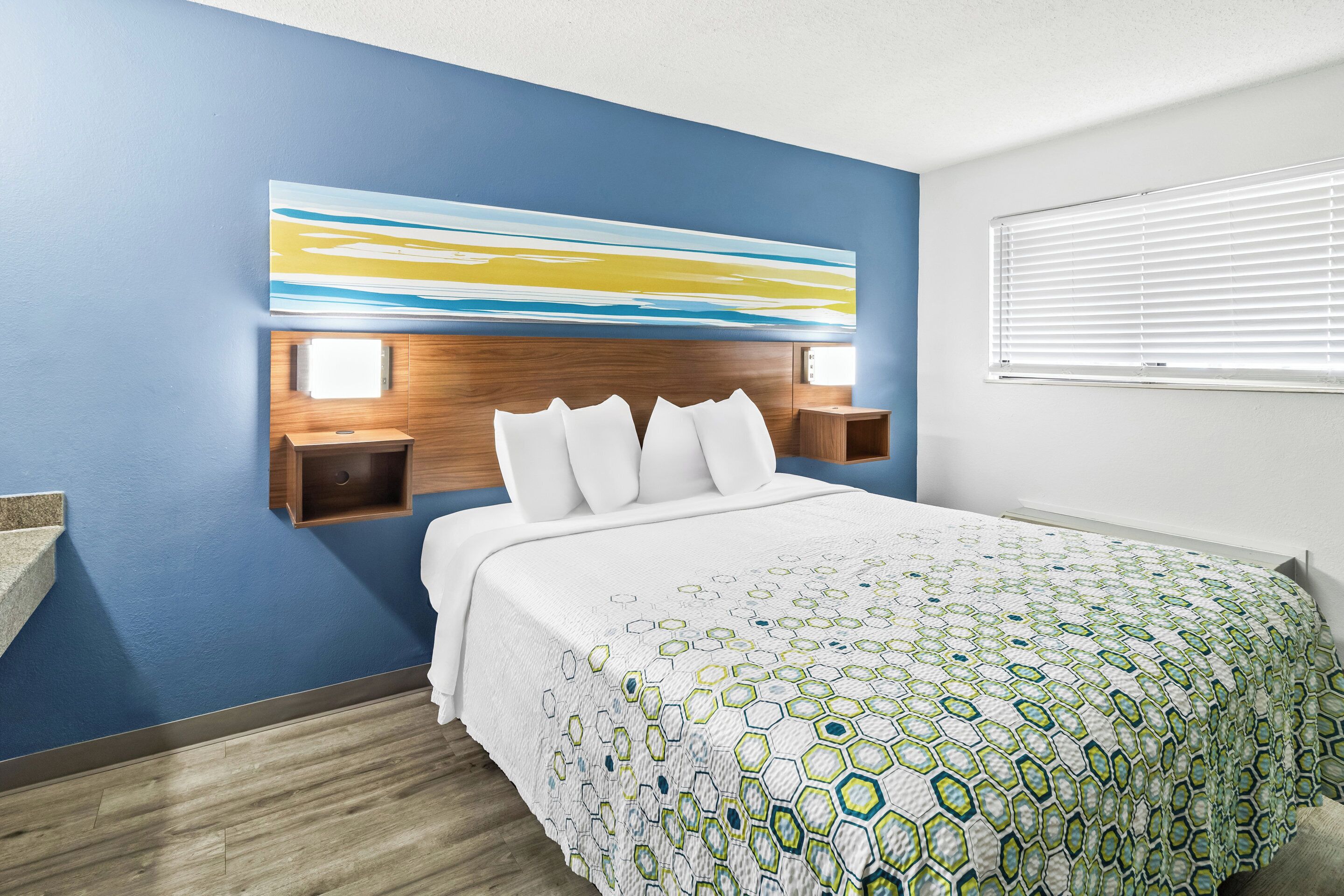 A hotel room with a bed, a nightstand, a wall-mounted lamp, and a window with blinds.
