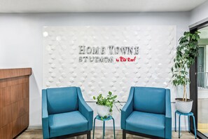 A reception area with a white textured wall, two blue chairs, a wooden reception desk, and a potted plant. - HomeTowne Studios by Red Roof Florence, KY-Cincinnati Airport (Florence)