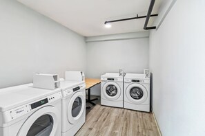 A laundry room with two commercial duty dryers and two front-loading washing machines. - HomeTowne Studios by Red Roof Florence, KY-Cincinnati Airport (Florence)