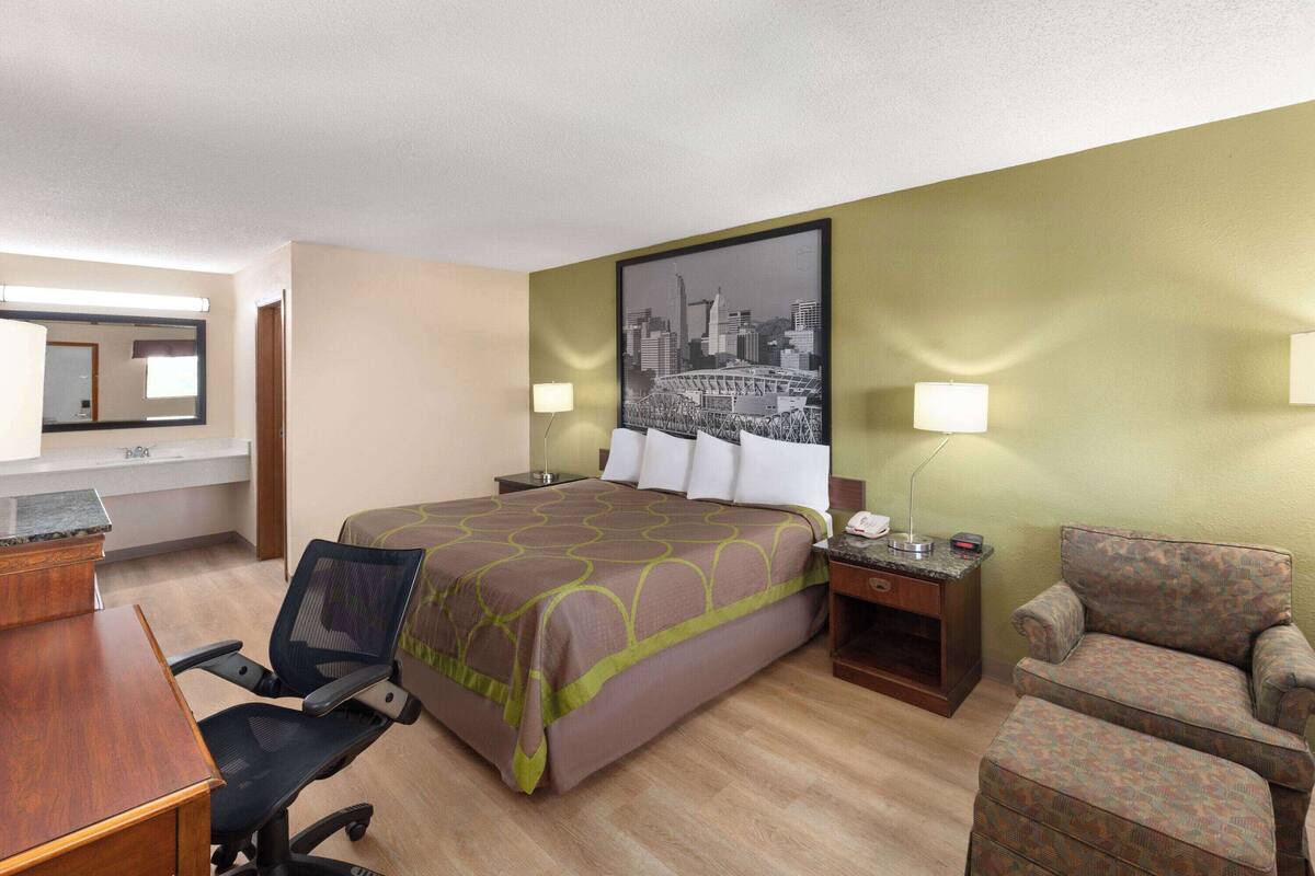A hotel room with a bed, a desk, a chair, a lamp, and a cityscape painting on the wall.