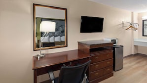 A hotel room with a wooden desk, a chair, a flat-screen TV, a refrigerator, and a mirror reflecting a cityscape. - Super 8 by Wyndham Fort Mitchell Cincinnati Area (Fort Mitchell)