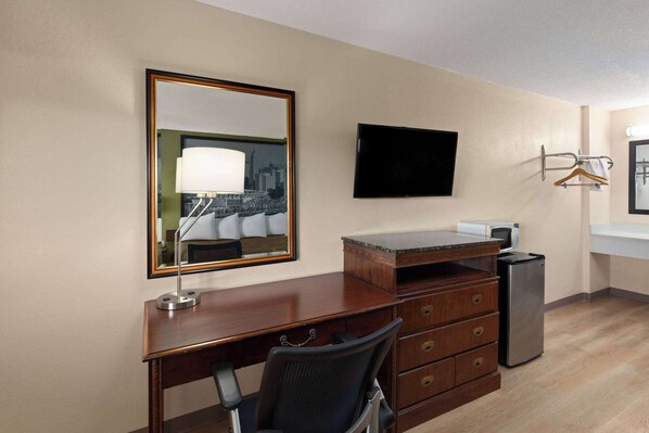 A hotel room with a wooden desk, a chair, a flat-screen TV, a refrigerator, and a mirror reflecting a cityscape.