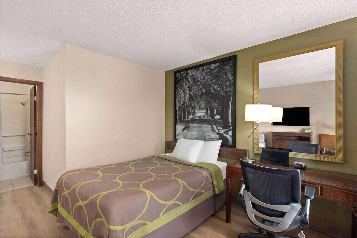A hotel room with a bed, a desk with a chair, a large mirror, and a framed picture on the wall.