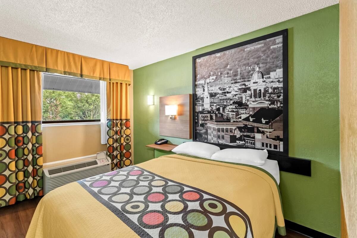 A hotel room with a bed, a window with curtains, a wall-mounted lamp, and a large cityscape poster.