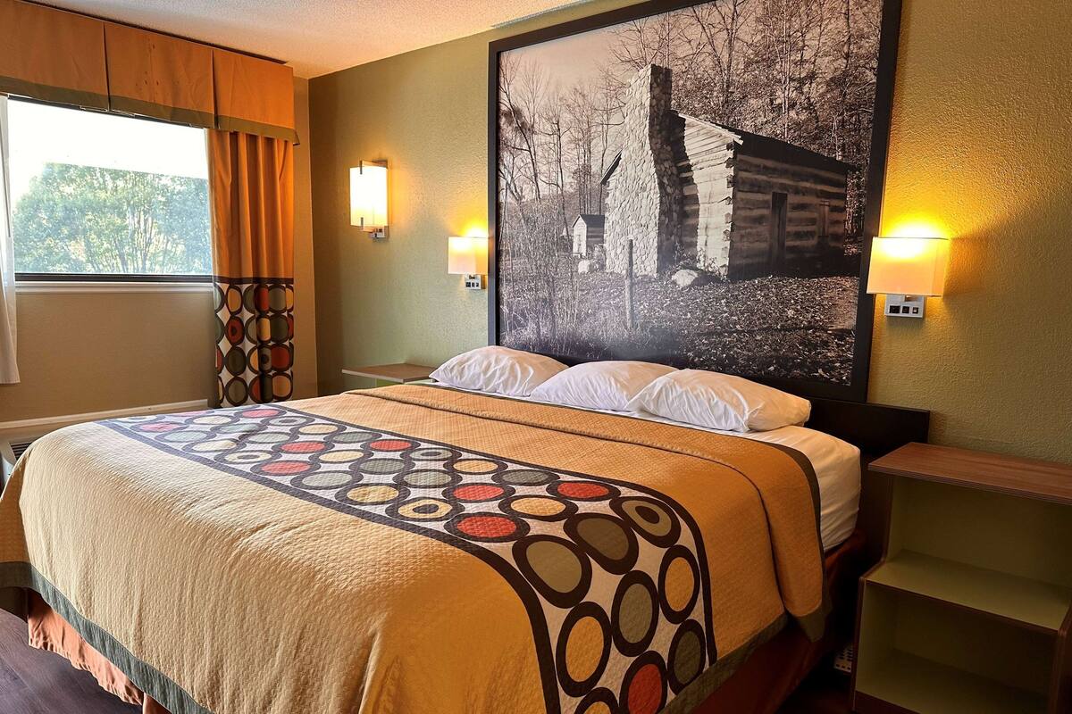 A hotel room with a bed, a large framed picture on the wall, and two wall-mounted lamps.