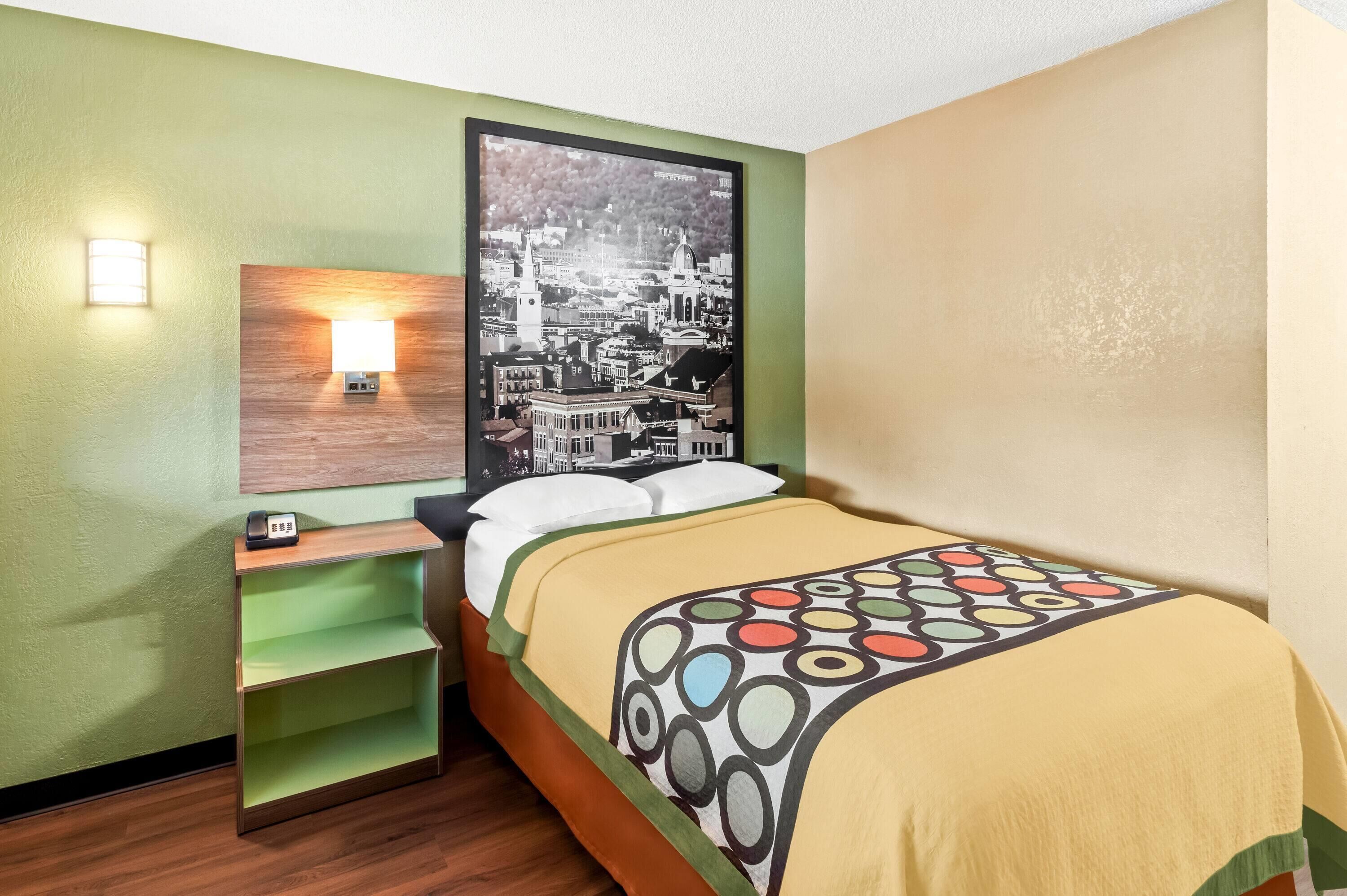 A hotel room with a bed, a nightstand, a wall-mounted lamp, and a cityscape artwork.