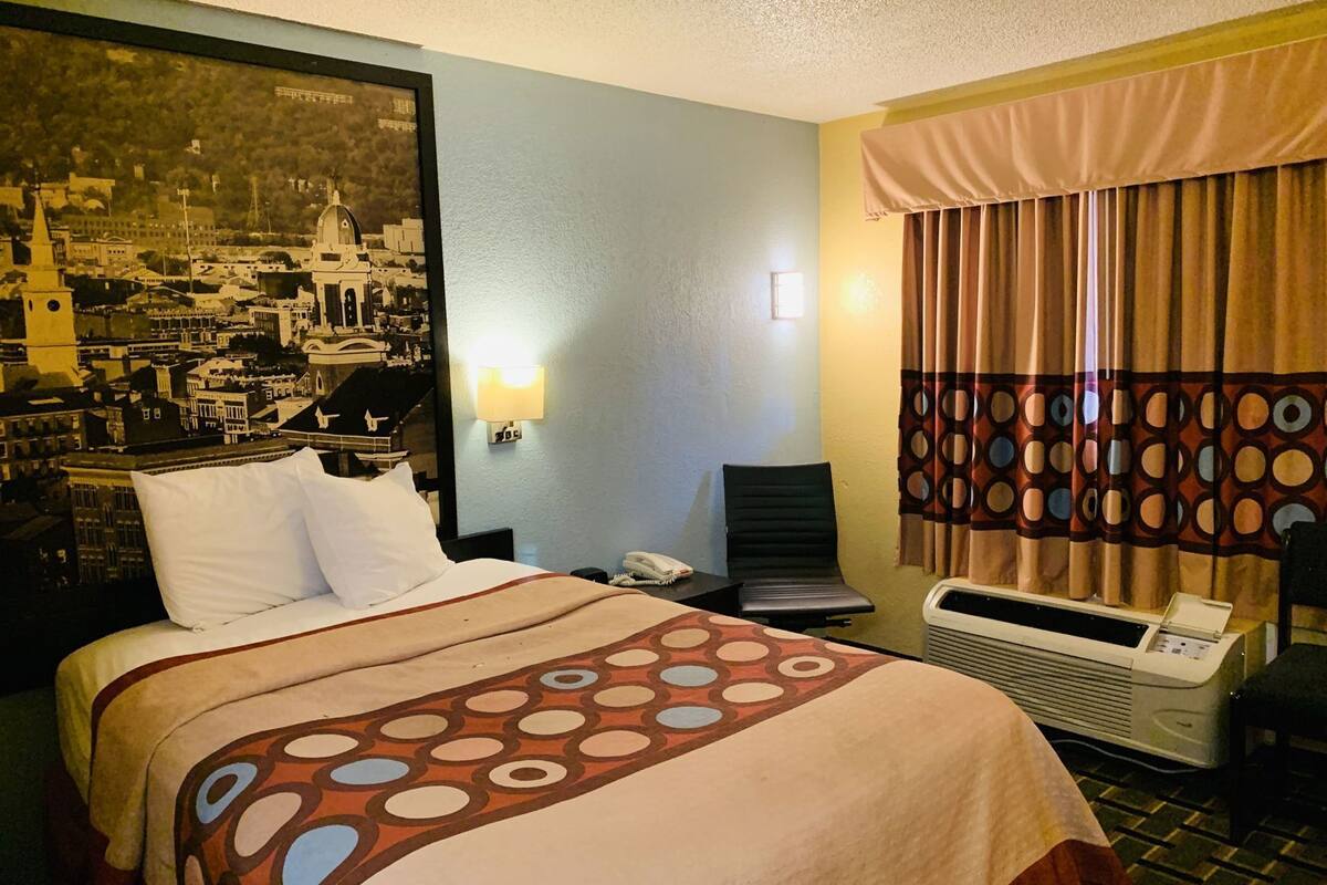A hotel room with a bed, a desk, a chair, a wall-mounted phone, a heater, and a cityscape painting.