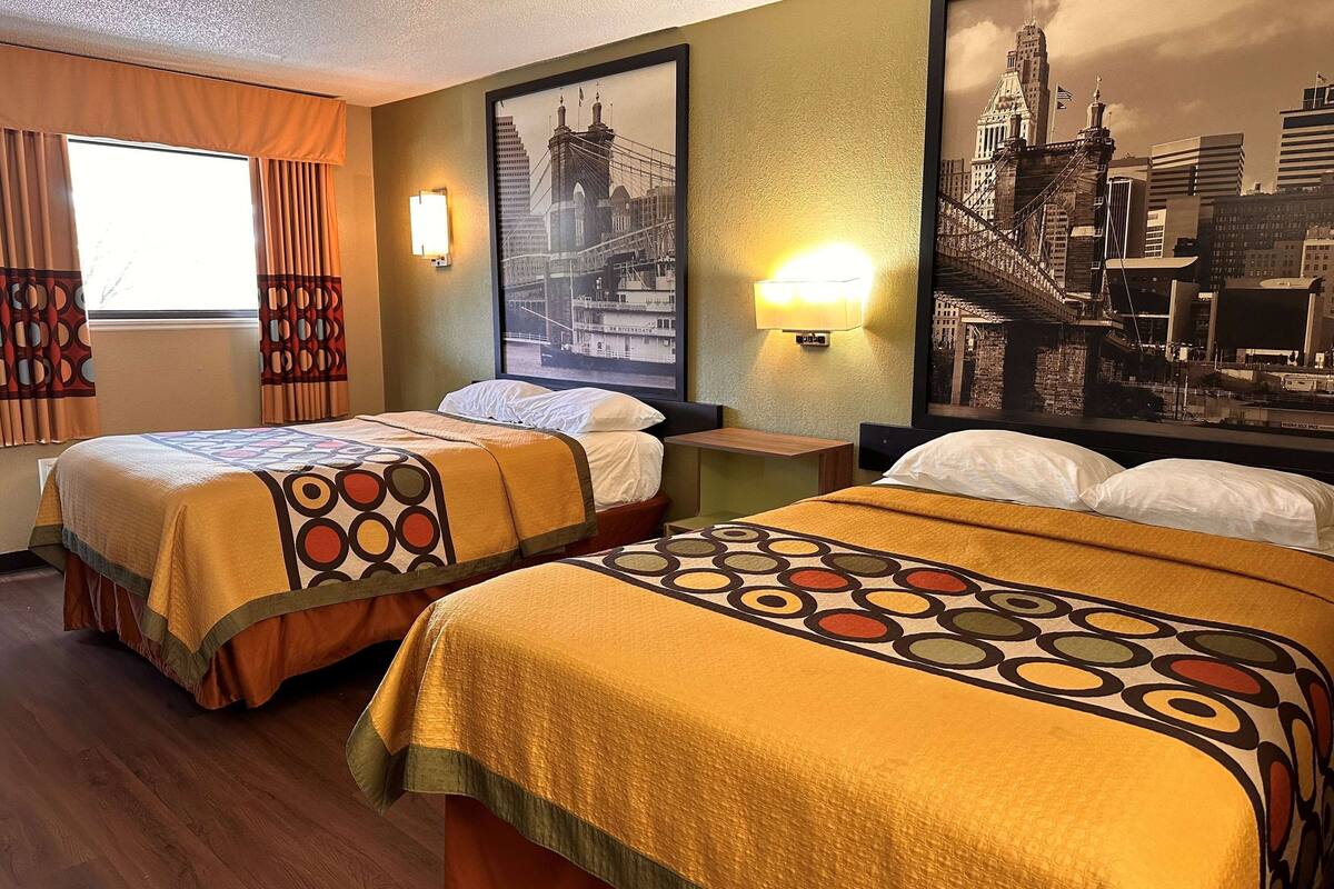 A hotel room with two beds, each with a patterned bedspread, a large cityscape painting on the wall, and a window with curtains.